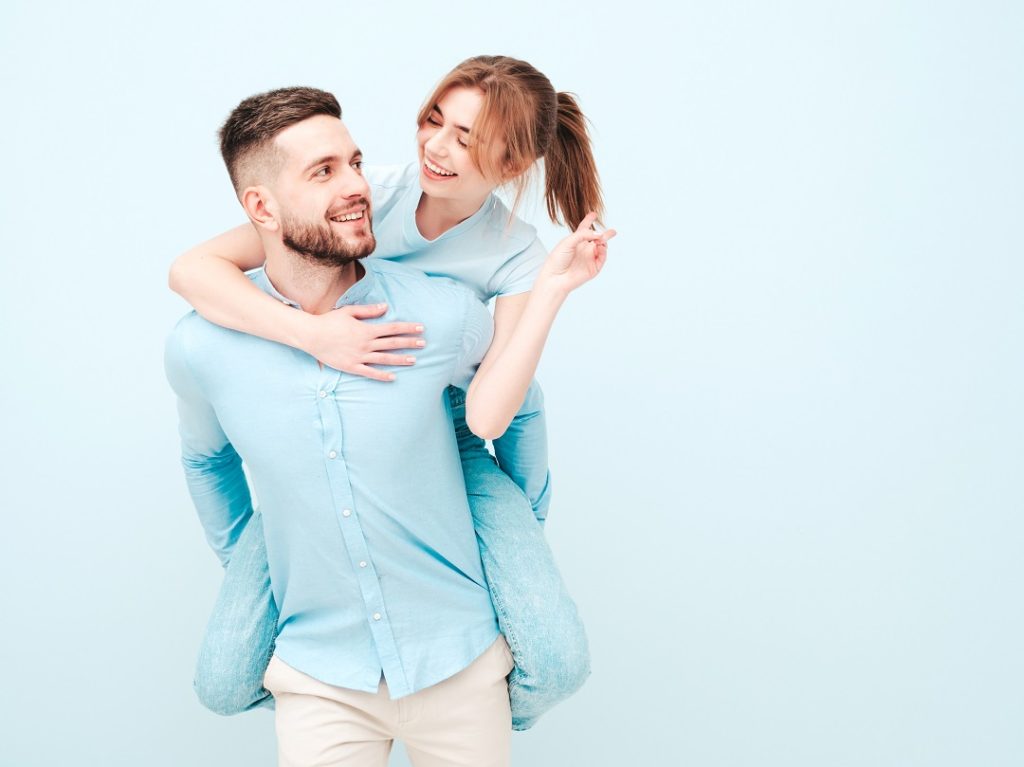 Smiling beautiful woman and her handsome boyfriend.Happy cheerful family having tender moments near light blue wall in studio.Pure models hugging.Embracing each other.Male gives piggyback riding